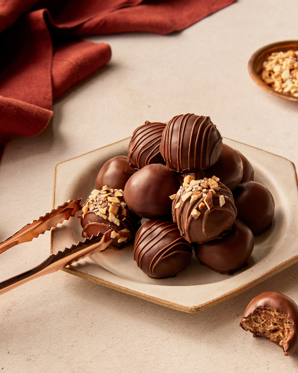 A pile of chocolate-dipped cookie dough bites on a hexagonal plate, being served with small tongs.