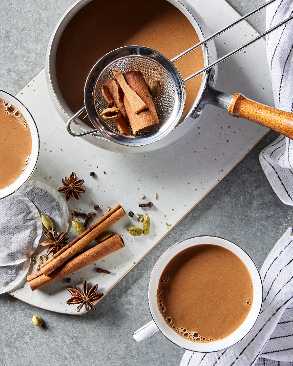 Top view of two tea cups of masala chai and a pot of chai with a strainer full of whole spices, shown with tea bags and assorted warm spices on a cutting board.