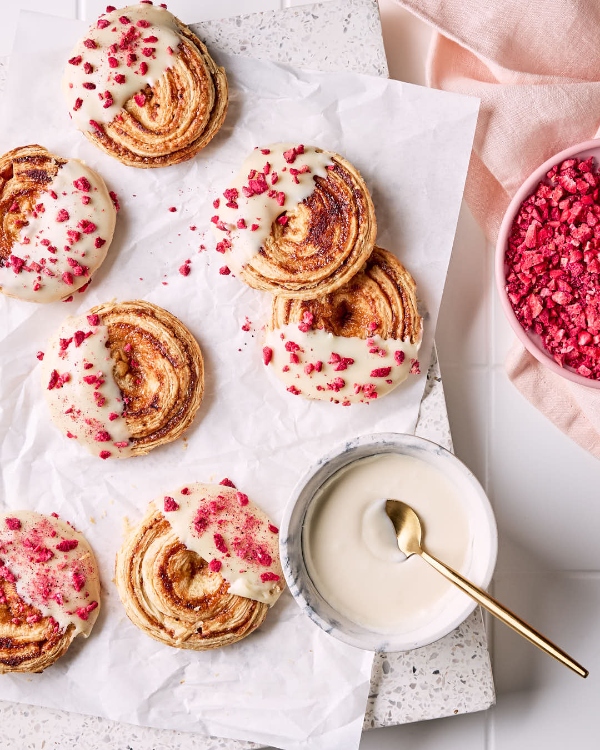 Raspberry puff pastry cookies iced with white chocolate and sprinkled with freeze-dried raspberries, a bowl of crushed raspberries, and a bowl with white chocolate dip with a gold spoon on parchment paper shown on a marble cutting board on a white tile countertop.