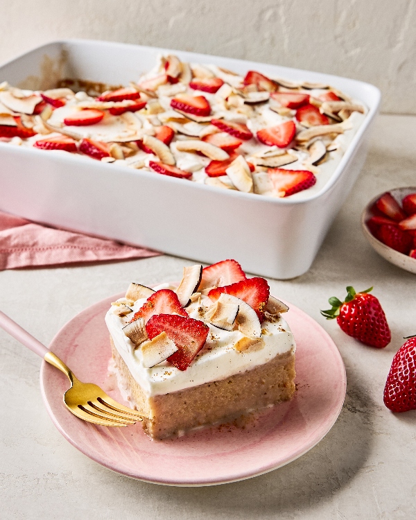Strawberry-Coconut Tres Leches cake slice on a pink plate with a gold fork, with the remaining cake in a white ceramic baking dish and fresh strawberries nearby, set on a textured grey surface with a pink linen napkin.