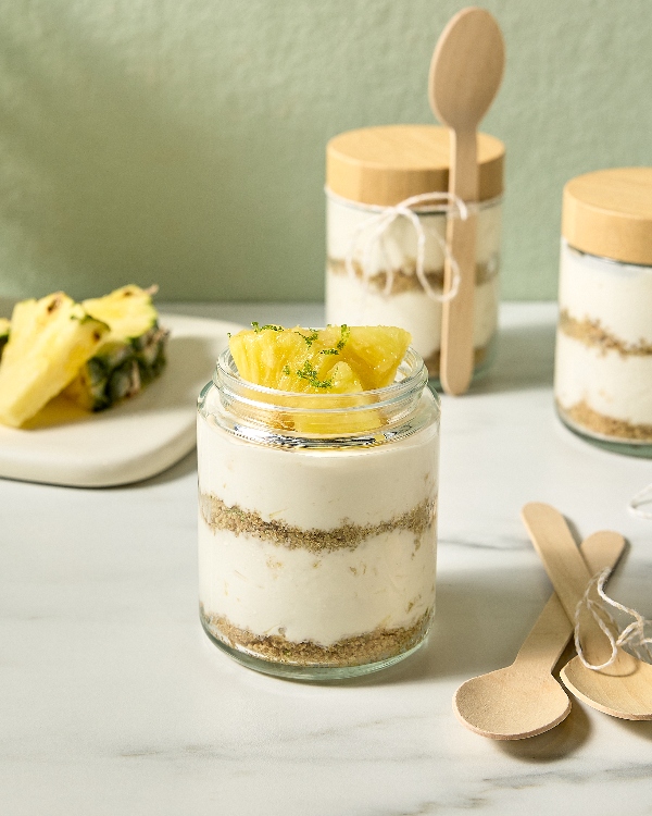 Several jars of single-serving pineapple cheesecake arranged on a white marble surface. Each jar features layers of creamy cheesecake and crumbly graham cracker crust, topped with fresh pineapple chunks and a sprinkle of lime zest. Wooden spoons are tied with string to the jars and a plate with extra pineapple pieces is visible in the background.