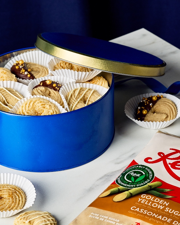 Gift tin filled with piped butter cookies, displayed in white paper liners. The cookies feature a variety of decorations, including chocolate-dipped with gold sprinkles, white chocolate drizzle, and golden sugar. A bag of golden yellow sugar is partially visible on a marble countertop, enhancing the baking theme.