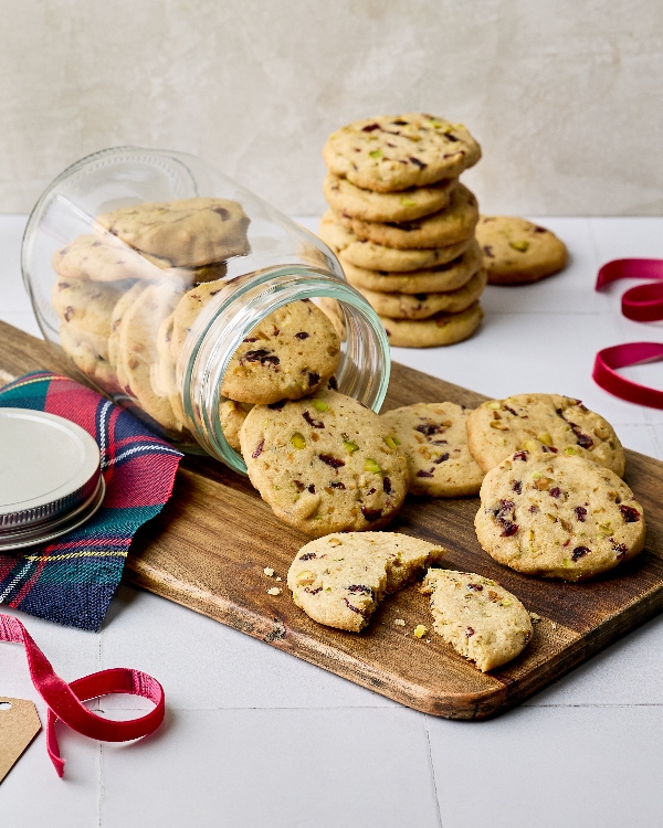 Cranberry pistachio cookies spilling from a glass jar onto a wooden board. A stack of cookies and a bag of baking ingredients are visible in the background. A red and green plaid napkin, along with a ribbon and gift tag, suggest a holiday gift-giving theme. One cookie is broken in half, showing a soft interior with chunks of cranberries and pistachios