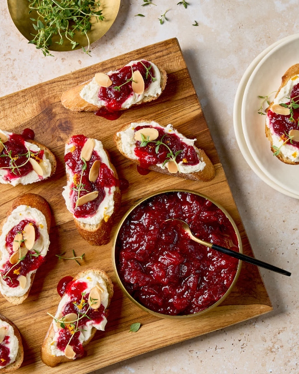 A wooden board filled with crostini topped with whipped feta cheese, cranberry chutney, slivered almonds, and fresh thyme. A small bowl of cranberry chutney is also on the board.