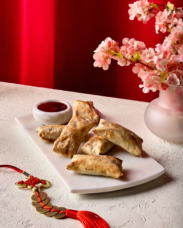 Golden apple spring rolls served on a white marble platter with a small bowl of red dipping sauce. The setup is accented by pink blossoms in a vase and a traditional red Chinese knot with gold coins, set against a vibrant red background.