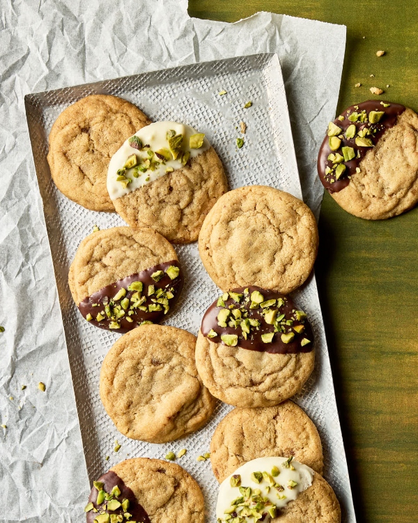 Chocolate chipless cookies arranged on a textured metal tray sitting on parchment paper. Some cookies are plain, while others are half-dipped in dark or white chocolate and sprinkled with chopped pistachios. A single decorated cookie rests on the green wooden surface beside the tray, with scattered pistachio pieces.