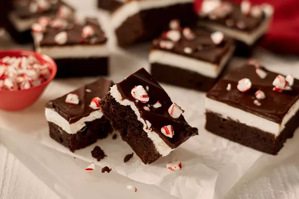 A close-up of chocolate mint brownies with a layer of mint cream and ganache top, each sprinkled with candy cane pieces, shown on wax paper on a white surface. 