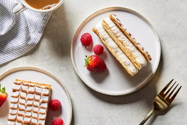 Two pieces of Mille-Feuille cake on plates, shown with berries, a cup of coffee and a gold fork.