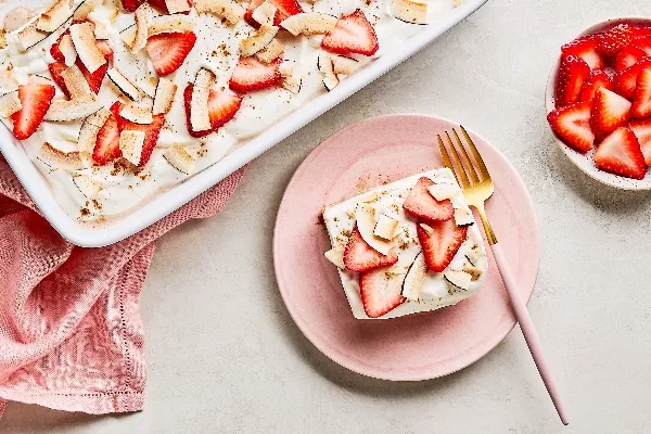 A single serving of Strawberry-Coconut Tres Leches cake on a pink plate with a gold fork, alongside a white baking dish of the same cake and a small bowl of sliced strawberries, all on a grey countertop with a pink cloth napkin