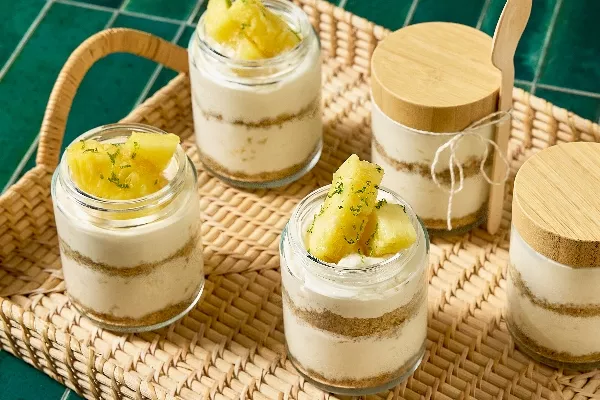  A wicker tray set against a green tiled background holds several jars of single-serving pineapple cheesecake. Each jar features layers of creamy cheesecake and crumbly graham cracker crust, topped with fresh pineapple chunks and a sprinkle of lime zest. Two of the jars have wooden lids, and one has a bamboo spoon tied to it.