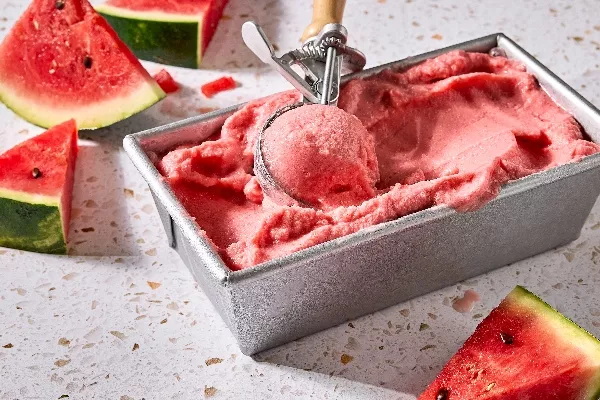 A metal container filled with creamy watermelon sherbet, being scooped with a vintage ice cream scooper. Fresh watermelon slices are scattered around the container on a light-coloured limestone countertop.