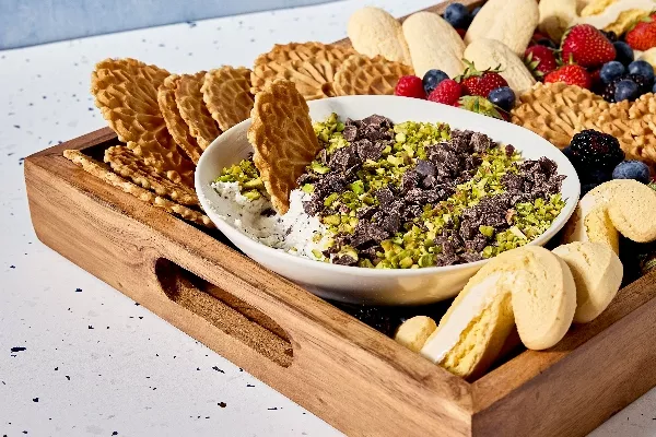 A wooden tray filled with an assortment of Italian cookies and fresh strawberries, blueberries, and blackberries surrounding a bowl of cannoli dip. The dip is topped with chopped pistachios and chocolate chunks and has a Pizzelle cookie dipped in.