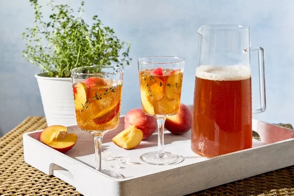 A white tray on a wicker table holds two stemmed glasses, a pitcher of peach-thyme sweet tea, and whole and cut fresh peaches. The drinks are garnished with fresh peach slices and sprigs of thyme, and a potted thyme plant is visible in the background.