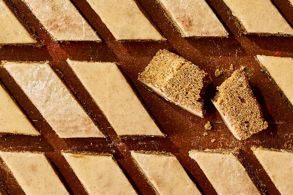 Rows of spiced cake bars cut into diamond shapes, topped with a smooth glaze. One bar is broken in half, revealing a moist and crumbly interior. The warm brown background looks rustic and cozy