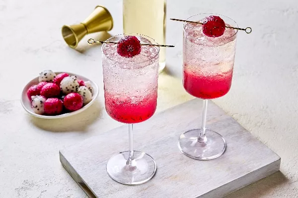 Two glasses of vibrant dragon fruit spritzer served over ice, with a pink gradient from the fruit at the bottom. Each glass is garnished with a skewered dragon fruit ball. A plate of red and white dragon fruit balls, a gold jigger, and a bottle are visible in the background.