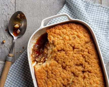 pudding chomeur in a dish with a scoop missing, the dish is sitting on a blue checkered napkin