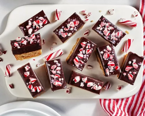 Golden cookie bars topped with chocolate ganache and peppermint stick pieces on a white tray with a red and white striped cloth