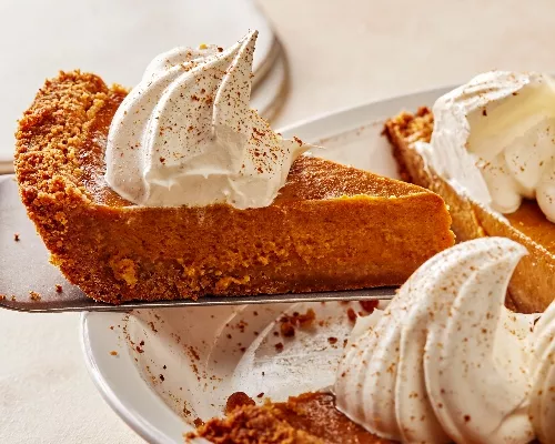 Removing a slice of pumpkin pie with whipped cream and ground cinnamon from a white pie plate with a pie lifter